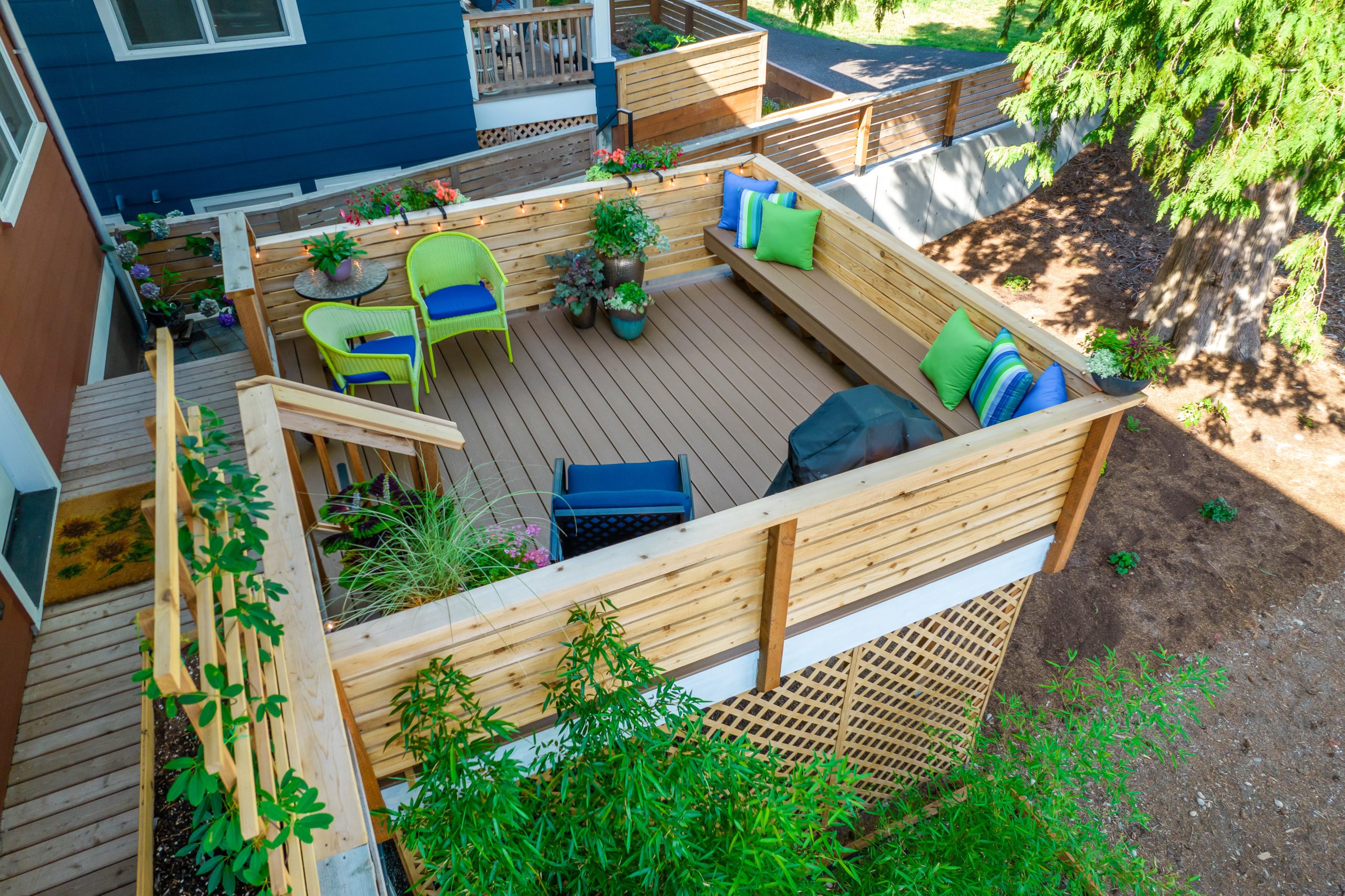 Outdoor deck patio made out of wood surrounded by trees designed with green and blue pillows and other decor