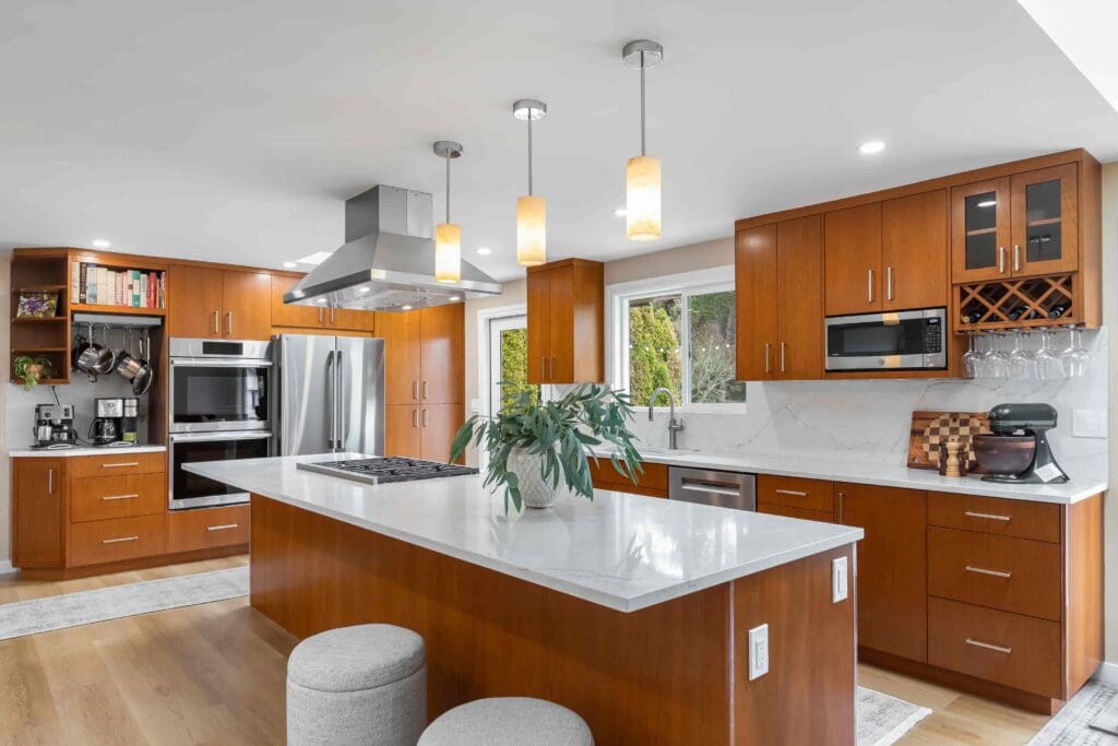11114 34th Pl SW, Seattle WA_0001 Modern kitchen with custom cherry cabinets, stainless steel appliances, a countertop island with two stools, pendant lights, and a plant centerpiece; large windows let in natural light.