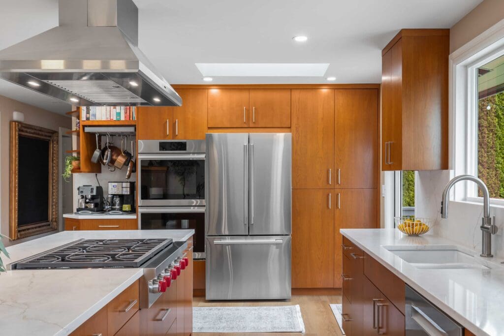 11114 34th Pl SW, Seattle WA_0003 Modern kitchen with stainless steel appliances, custom cherry cabinets, a large refrigerator, an oven, a stove with a hood, a coffee station, white countertops, a sink by the window, and a bowl of bananas on the counter.