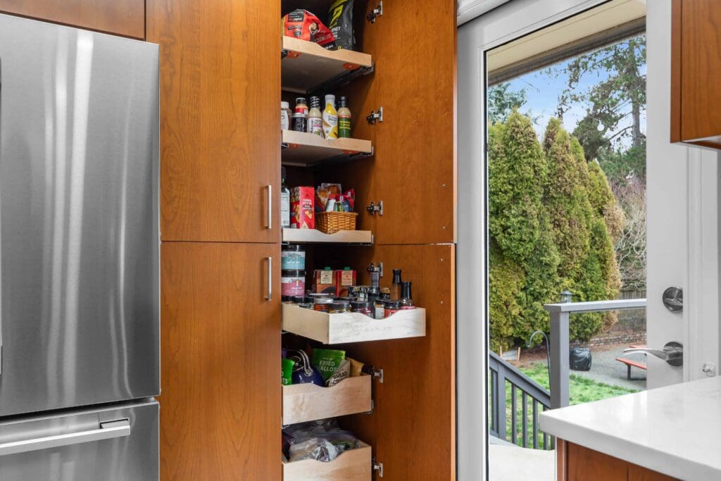 11114 34th Pl SW, Seattle WA_0005 A modern kitchen features custom cherry cabinets, including a tall wooden pantry cupboard open to reveal organized shelves of food items, next to a stainless steel refrigerator and a glass door leading to a backyard with greenery.
