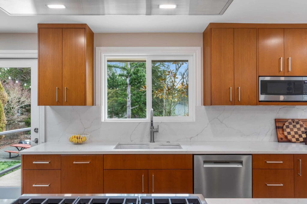 11114 34th Pl SW, Seattle WA_0007 Modern kitchen with custom cherry cabinets, white countertops and backsplash, stainless steel appliances, a large window above the sink showing green trees outside, and a bowl of lemons on the counter.
