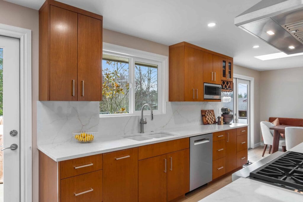 11114 34th Pl SW, Seattle WA_0008 Modern kitchen with custom cherry cabinets, white countertops, stainless steel appliances, a sink below a window, and a dining area in the background. A bowl of lemons sits on the counter near the sink.
