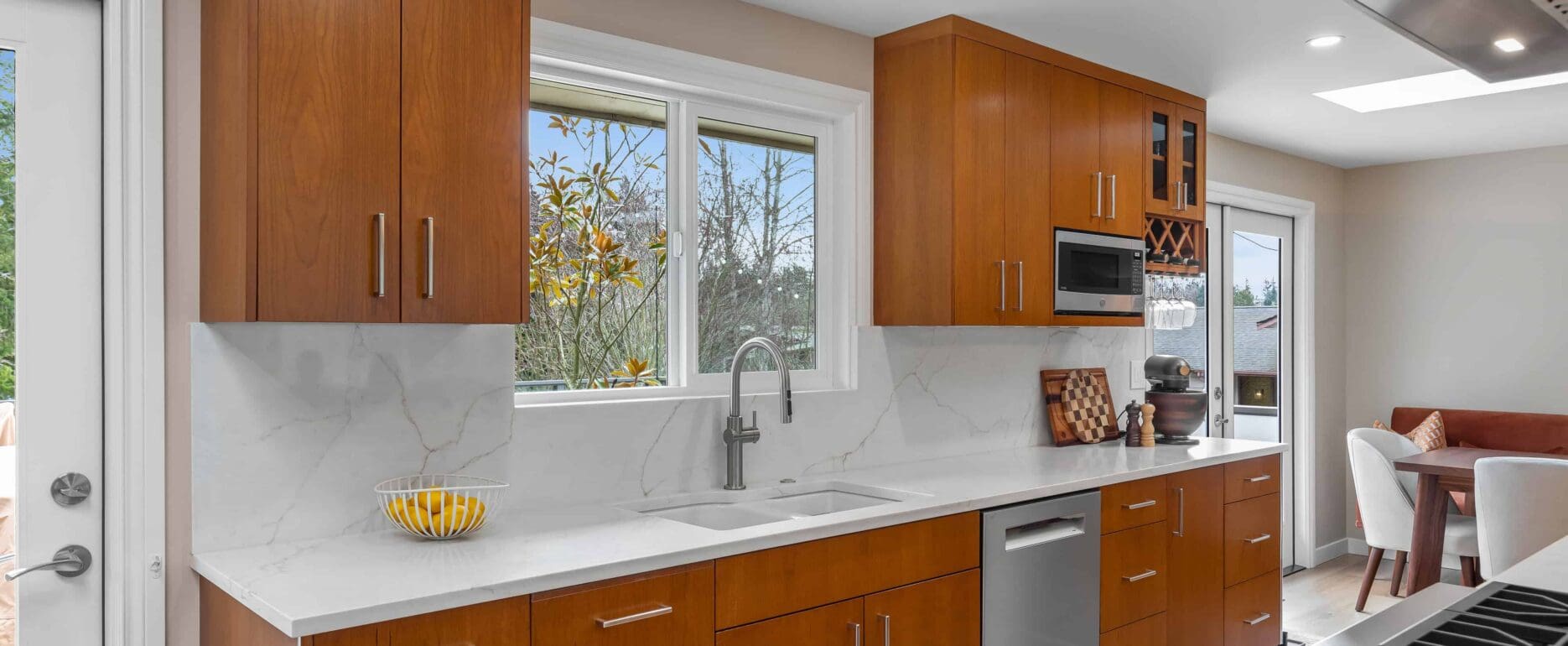 Modern kitchen with custom cherry cabinets, white countertops, stainless steel appliances, a sink below a window, and a dining area in the background. A bowl of lemons sits on the counter near the sink.