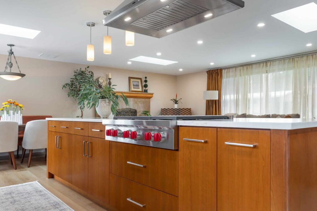 11114 34th Pl SW, Seattle WA_0011 Modern kitchen with custom cherry cabinets, a large island featuring a stove with red knobs, pendant lighting, and an open layout leading to a dining area with a table, chairs, and large windows with sheer curtains.