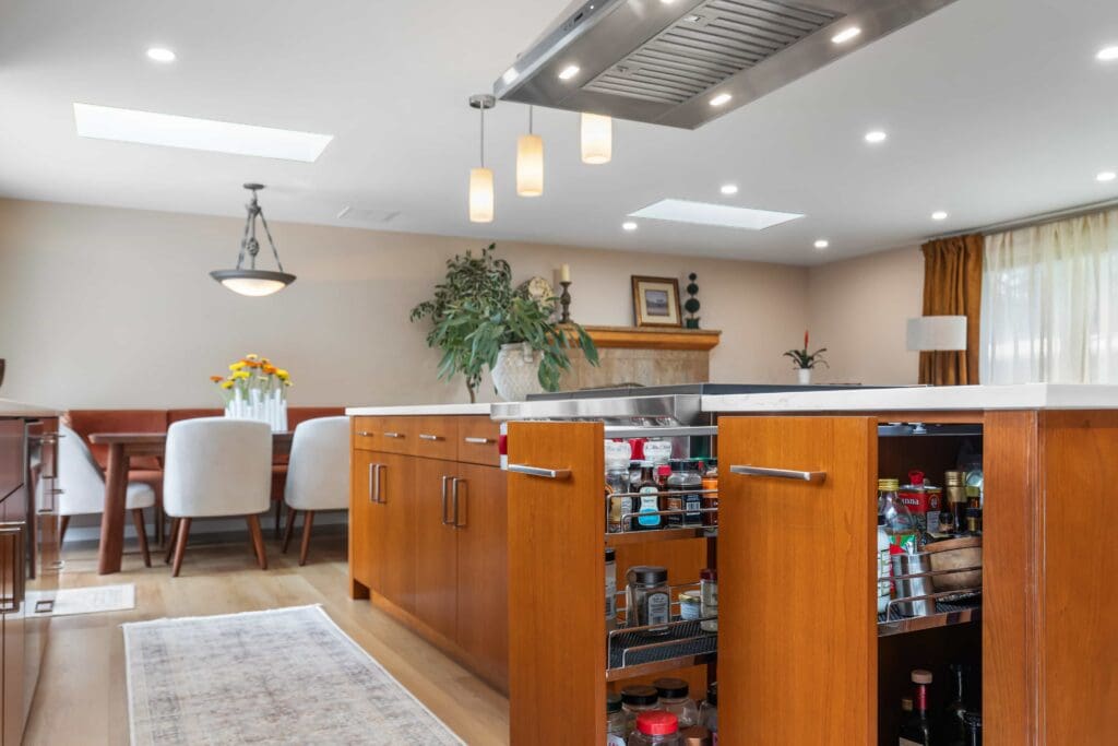 11114 34th Pl SW, Seattle WA_0012 Modern kitchen with custom cherry cabinets and an island featuring open spice drawers. Pendant lights hang above the island, while a dining area with chairs and a table is visible in the background, along with large windows and plants.