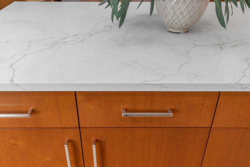 11114 34th Pl SW, Seattle WA_0014 Close-up of a white countertop with subtle gray veining, atop custom cherry cabinets with silver handles. A decorative vase with green leaves is partially visible on the counter.