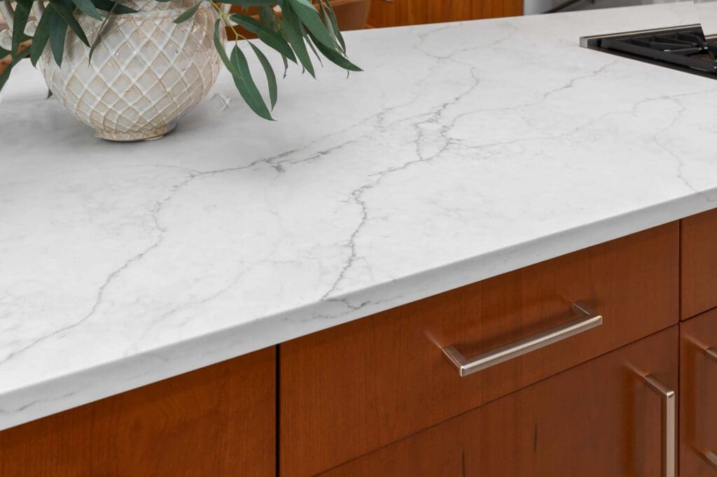 11114 34th Pl SW, Seattle WA_0015 White countertop with subtle gray veining, set atop custom cherry cabinets with silver handles. A decorative plant in a textured ceramic pot sits on the counter, and part of a stovetop is visible on the right.