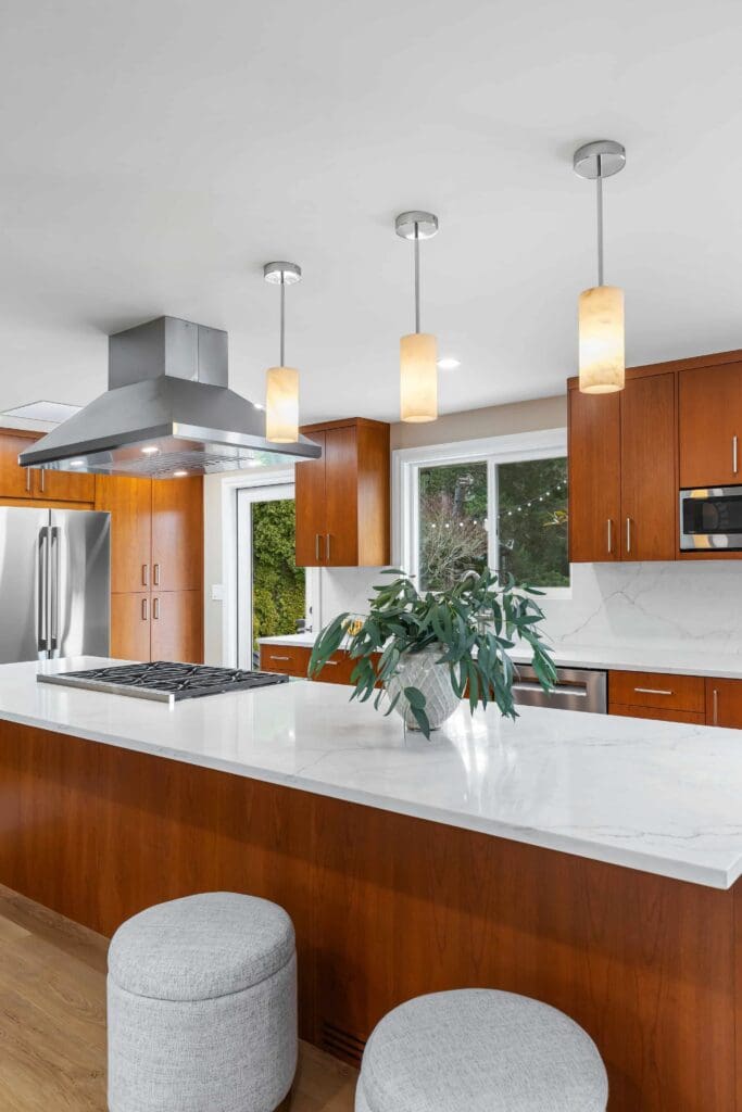 11114 34th Pl SW, Seattle WA_0018 Modern kitchen with custom cherry cabinets, white countertops, stainless steel appliances, three pendant lights, a range hood, a plant centerpiece on the island, and two round gray stools in the foreground.