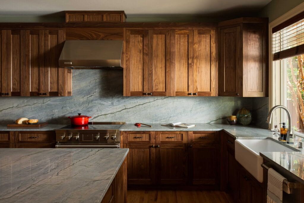 HAAS-Seattle-Kitchen-Remodel-3 (1) A modern kitchen featuring Seattle custom cabinets in dark wood, gray marble countertops and backsplash, a stainless steel stove with a red pot, farmhouse sink, and sunlight streaming through a window with wooden blinds.