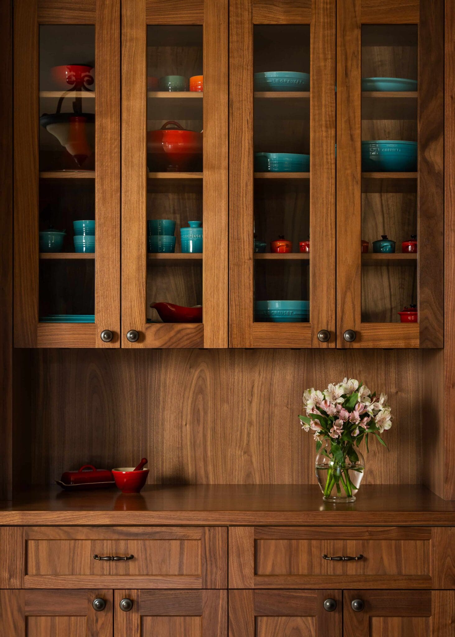 A wooden kitchen cabinet with glass doors, crafted by Seattle custom cabinets, displays colorful blue and red dishes. Below, a countertop holds a glass vase of light pink flowers and a small red dish. The cabinet has a warm, natural wood finish.