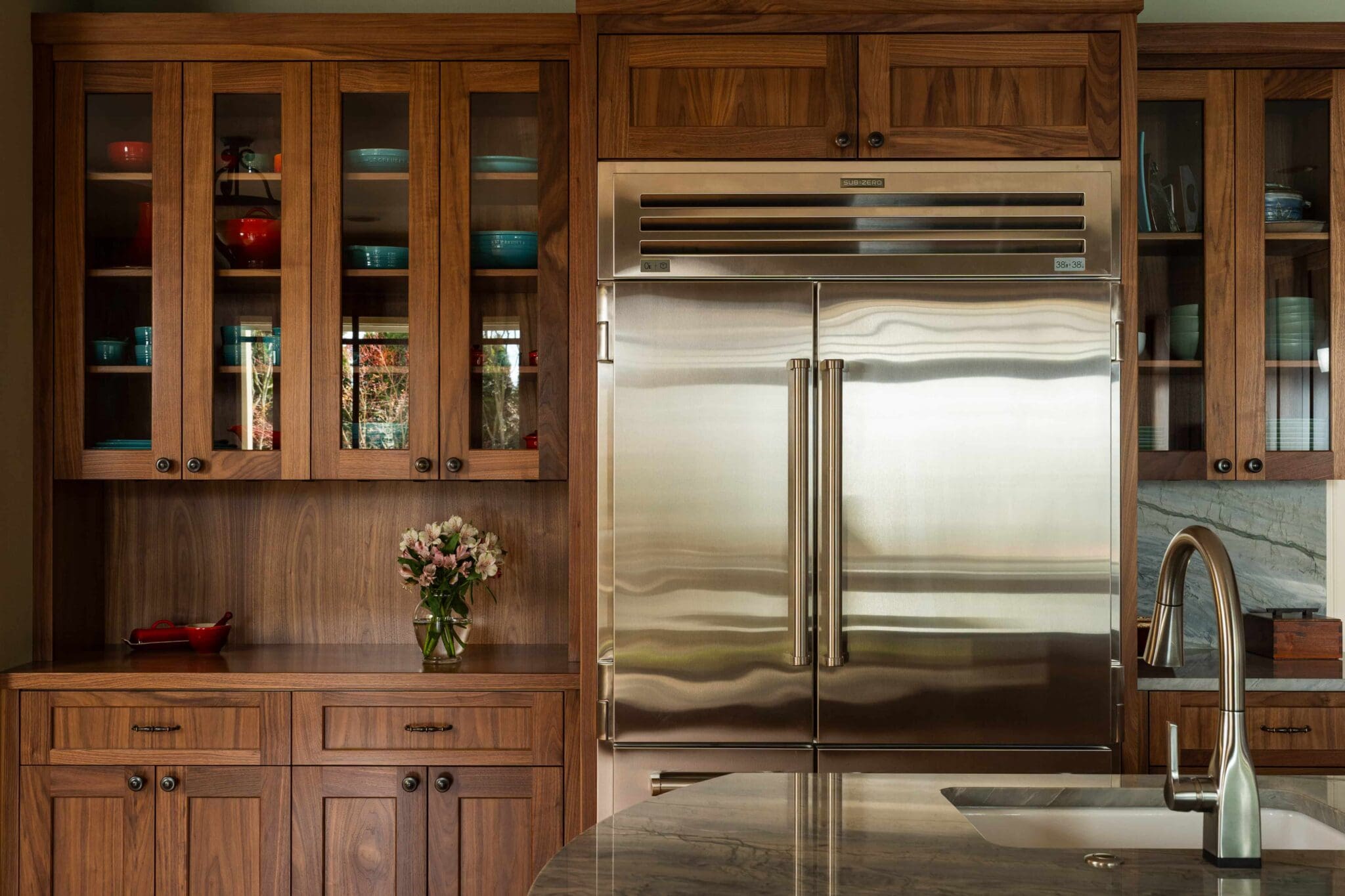 Modern kitchen with Seattle custom cabinets, a stainless steel double-door fridge, glass front cupboards displaying colorful dishes, a vase of flowers on the counter, and a sink with a silver faucet in the foreground.