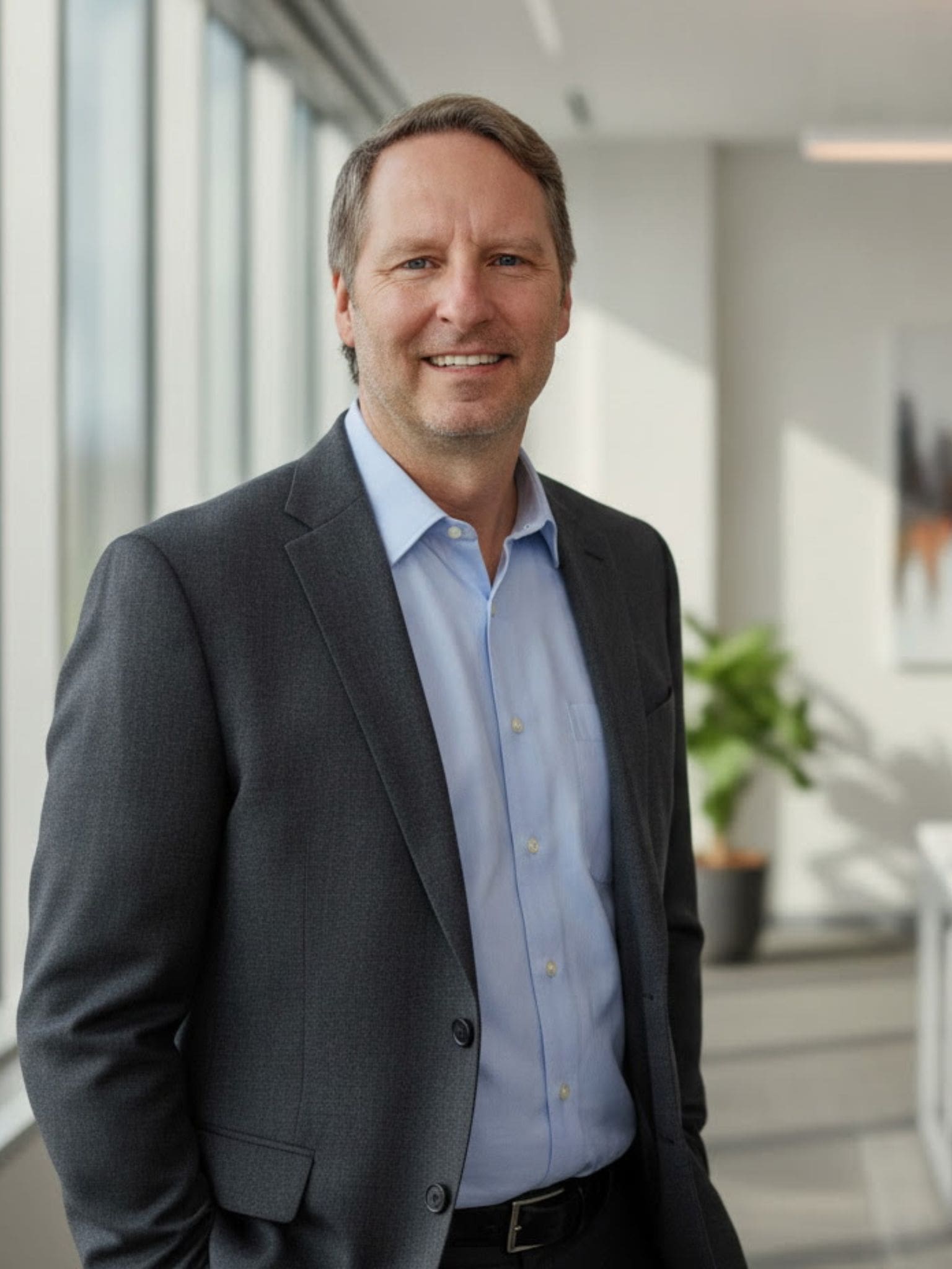 Wade Damey Wade in a suit smiles while standing in a bright office with large windows, a plant, and blurred artwork in the background.