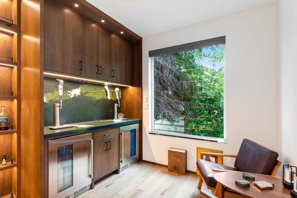 Custom Home Whiskey Bar 24 Modern kitchenette with wooden cabinets, two beverage taps, under-counter fridges, and a large window showing greenery. A custom home bar feel is created by the leather chair and small table with books and coasters in the corner.