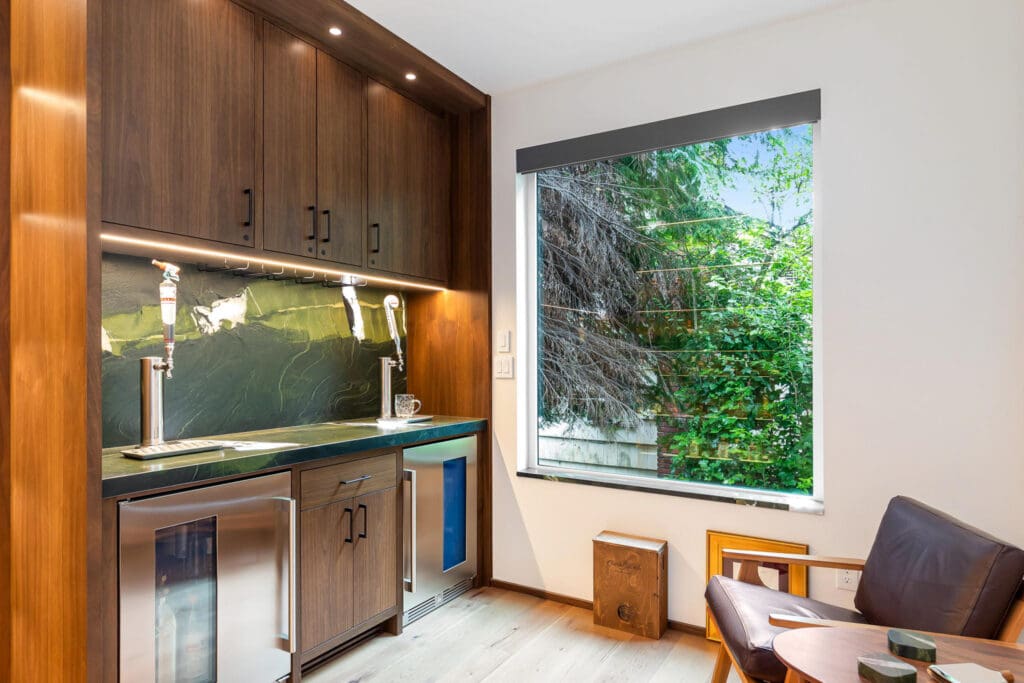 Custom Home Whiskey Bar 1 Small modern kitchenette with dark wood cabinets, a built-in mini fridge, stone countertop, and a large window overlooking green trees. A custom home bar is seamlessly integrated into the layout, while a leather chair and small table sit in the corner by the window.
