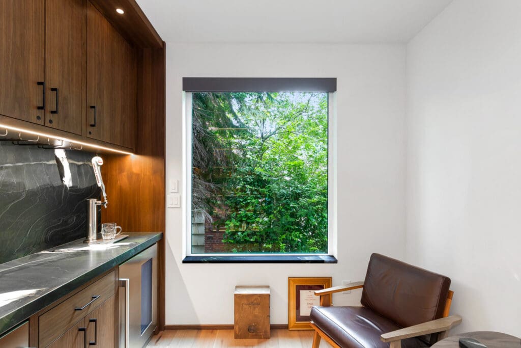 Custom Home Whiskey Bar 2 Modern room with dark wood cabinets, marble counter, and a small sink—perfect for a custom home bar. Large window shows green trees outside. A brown leather chair and framed items are near the window. Natural light brightens the space.