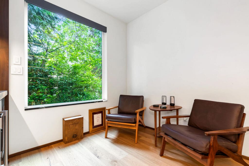 Custom Home Whiskey Bar 3 A cozy room with two wooden armchairs, a small round table, a cube-shaped wooden stool, a framed certificate, and a large window showing green trees outside—perfect for relaxing or entertaining guests near your custom home bar.