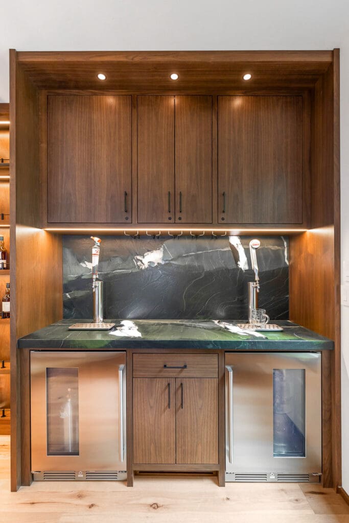 Custom Home Whiskey Bar 5 A modern custom home bar featuring dark wood cabinets, a sleek black marble backsplash, two beer taps, under-cabinet lighting, and two stainless steel mini fridges beneath a dark countertop.