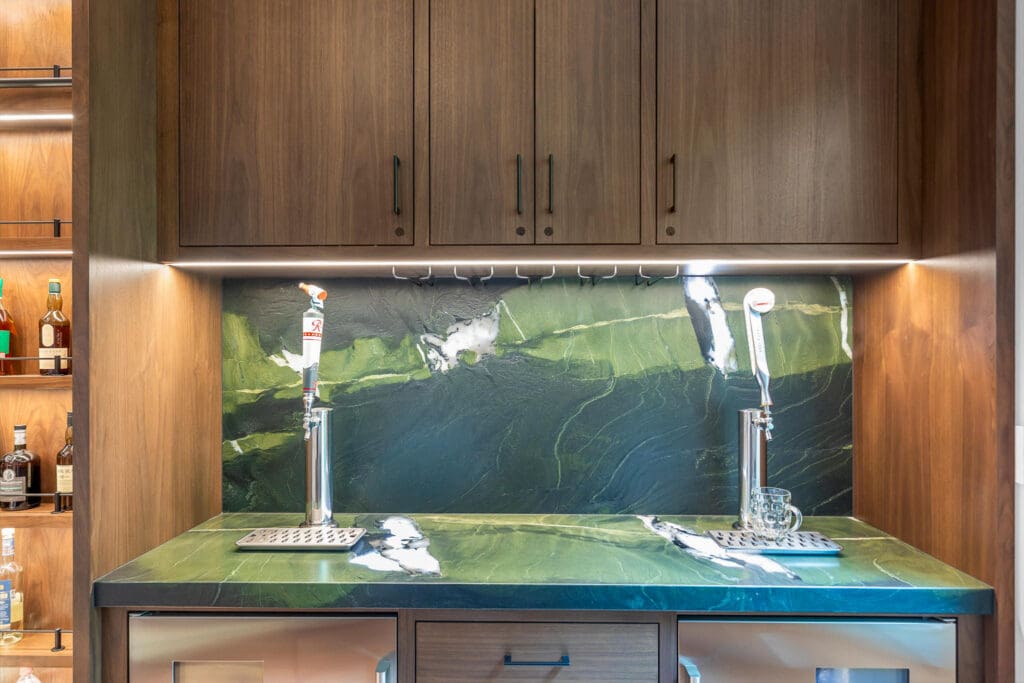 Custom Home Whiskey Bar 6 A custom home bar setup with a green marble countertop and backsplash, two beer taps, a glass, and wooden cabinets above. Bottles of liquor are visible on shelves to the left.