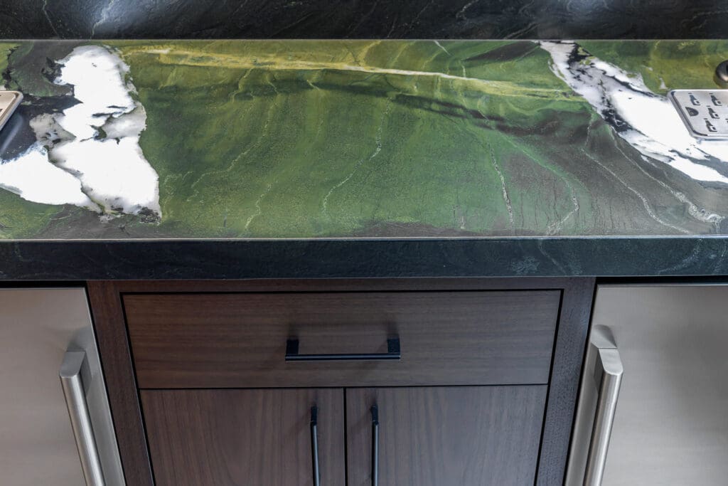 Custom Home Whiskey Bar 7 A close-up of a kitchen countertop with a green, black, and white marble pattern, above a dark wood cabinet with black and silver handles—perfect for elevating the look of a custom home bar.