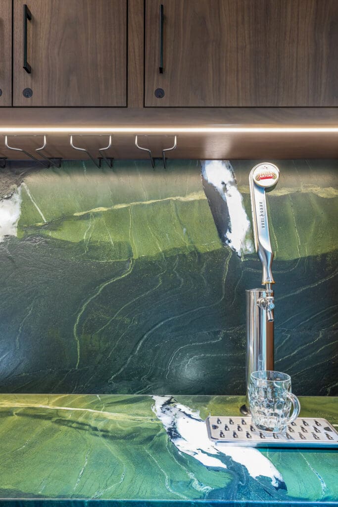 Custom Home Whiskey Bar 8 A close-up of a custom home bar setup on a kitchen counter with a green marble backsplash, a wooden cabinet above, a beer tap handle, a metal drip tray, and a clear glass mug under the tap.