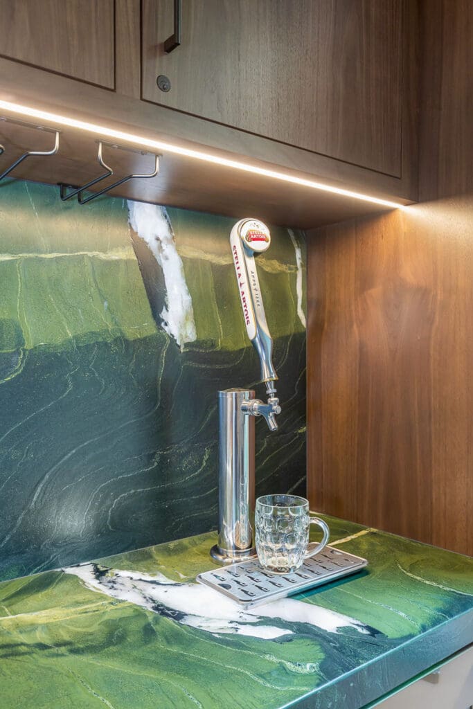 Custom Home Whiskey Bar 9 A custom home bar features a beer tap with a handle labeled Canadian Blond on a green marble countertop, with a glass beer mug beneath the tap, all set under wooden cabinets highlighted by under-cabinet lighting.