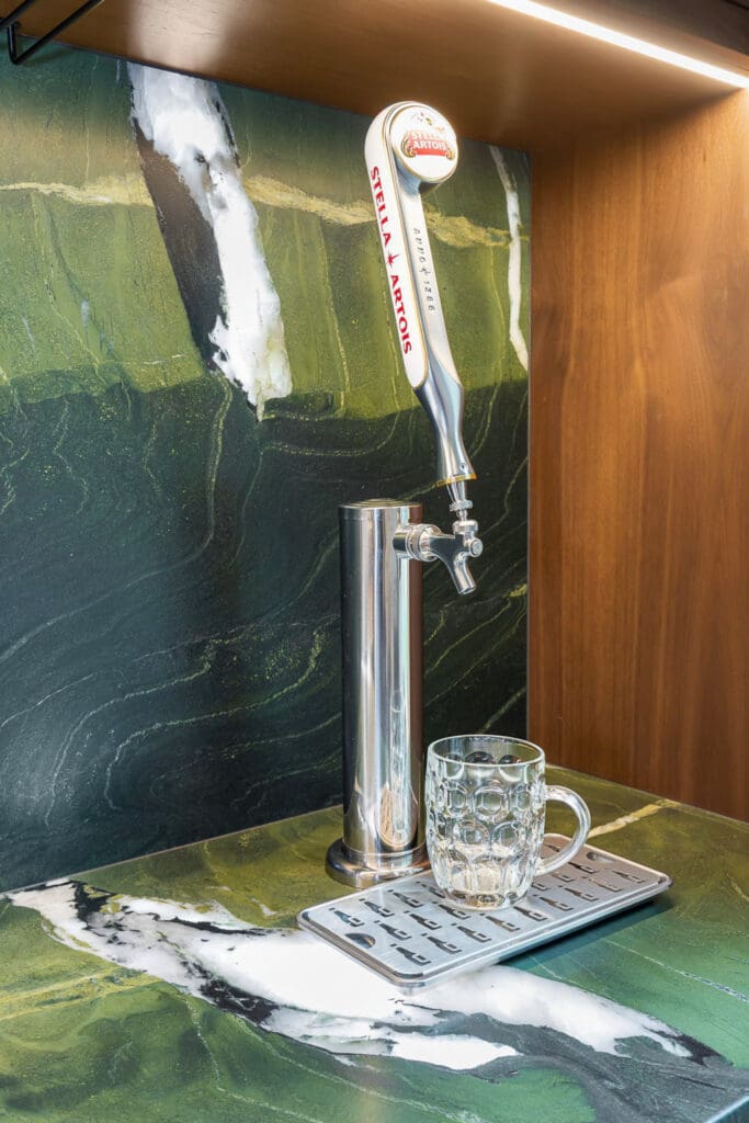 Custom Home Whiskey Bar 10 A beer tap labeled Stella Artois stands on a green marble countertop with a clear glass mug below it, ready for pouring—an inviting centerpiece for any custom home bar. The background is a matching green marble wall.