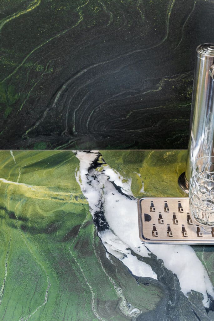 Custom Home Whiskey Bar 13 A green and black marbled countertop with white veins—perfect for a custom home bar—features a tall clear glass and a metallic drip tray placed on the right side of the surface.