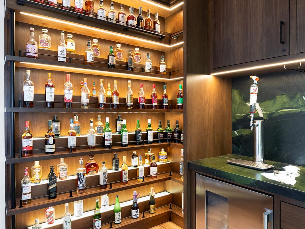 A custom home bar features wooden shelves displaying various liquor bottles, a small sink, under-cabinet lighting, a mini fridge, and elegant wood flooring.