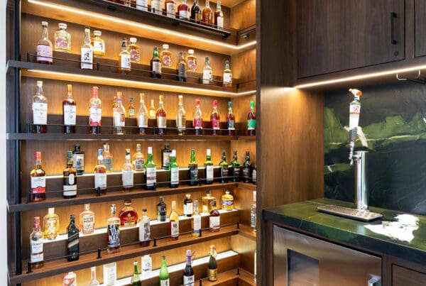 A custom home bar features wooden shelves displaying various liquor bottles, a small sink, under-cabinet lighting, a mini fridge, and elegant wood flooring.