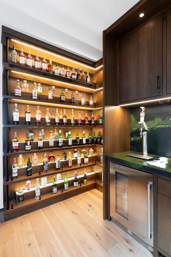Custom Home Whiskey Bar 16 A custom home bar features wooden shelves displaying various liquor bottles, a small sink, under-cabinet lighting, a mini fridge, and elegant wood flooring.