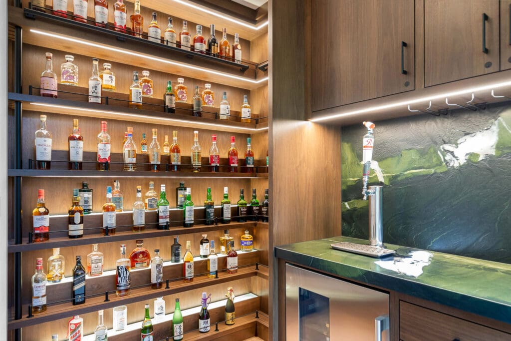 Custom Home Whiskey Bar 17 A custom home bar with shelves of assorted liquor bottles, a green marble countertop, mini fridge, and a beer tap. Wooden cabinets and under-shelf lighting create a modern, upscale look.