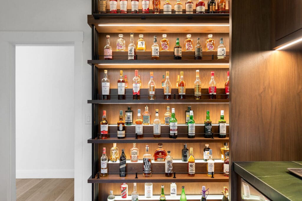 Custom Home Whiskey Bar 18 A well-lit custom home bar features shelves filled with various liquor bottles, arranged neatly against a wooden wall. To the left is a white doorway leading to a bright room with white walls and light wood floors.