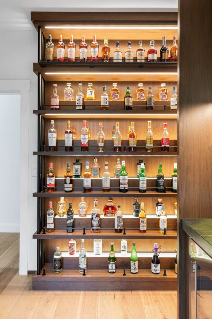 Custom Home Whiskey Bar 19 A wall-mounted wooden shelf in this custom home bar displays various bottles of liquor, including whiskey, vodka, gin, and sake, arranged neatly in rows with accent lighting highlighting the collection.