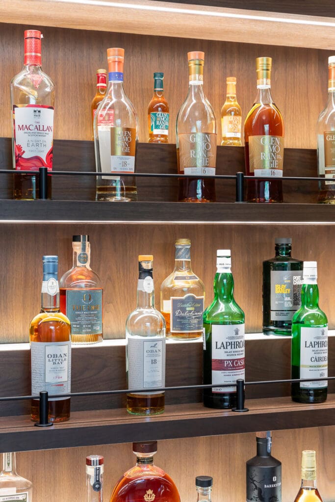 Custom Home Whiskey Bar 20 Bottles of various whiskey and liquor brands are displayed on wooden shelves in a custom home bar, arranged in rows with some recognizable labels and different shapes and colors of bottles visible.