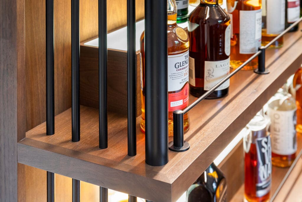 Custom Home Whiskey Bar 21 Bottles of whiskey and other spirits are arranged on wooden shelves with black metal bars in front, illuminated by built-in lighting, creating the perfect display for a custom home bar.