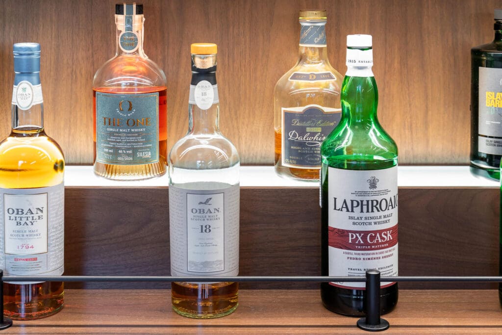 Custom Home Whiskey Bar 22 A display of various whiskey bottles, including Oban, The One, and Laphroaig PX Cask, arranged on a wooden shelf with a metal rail in front—perfect for adding a touch of sophistication to your custom home bar.