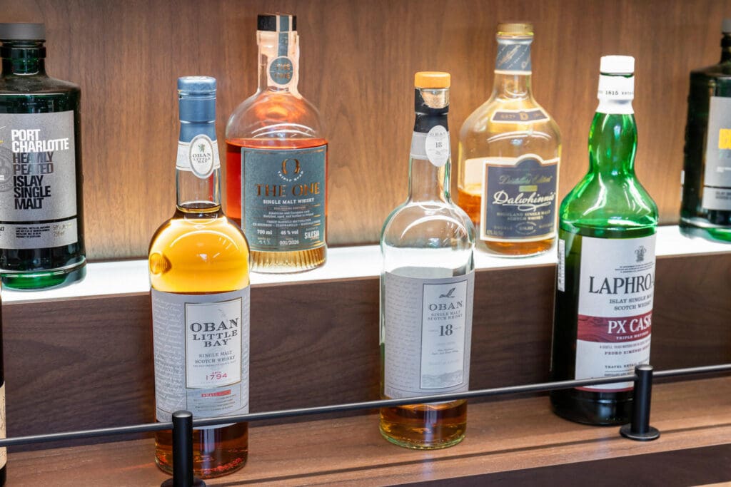Custom Home Whiskey Bar 23 A row of assorted whisky bottles is displayed on wooden shelves in a custom home bar, including brands like Oban, Laphroaig, and Dalwhinnie. The bottles are neatly arranged in a well-lit setting.