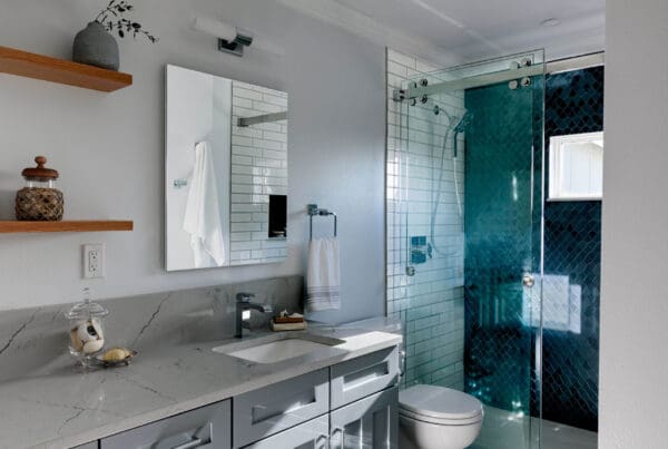 Modern West Seattle Craftsman Teens Bathroom with gray cabinets, marble countertop, wall mirror, glass shower with blue tiles, toilet, shelves with jars, and a towel hanging beside the sink.