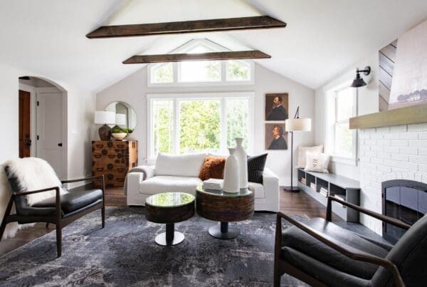 Bright living room with exposed wood beams, modern furniture, a white sofa, black armchairs, round coffee tables, a patterned rug, large windows—part of the stylish Greenwood Charmer Remodel.