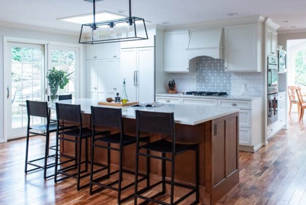 Modern kitchen with a large island, dark bar stools, white cabinetry, stainless appliances, wood floors, and subway tile backsplash. This Bellevue Kitchen Remodel features glass doors leading outside and a dining area in the background.