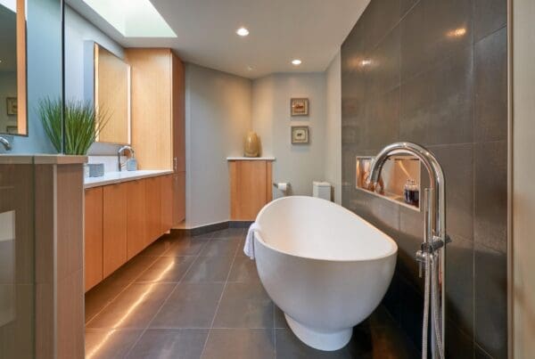 Modern bathroom in a West Seattle Contemporary Remodel featuring a white oval bathtub, freestanding faucet, dark tile walls and floors, light wood cabinets, a skylight, and minimalist decor.