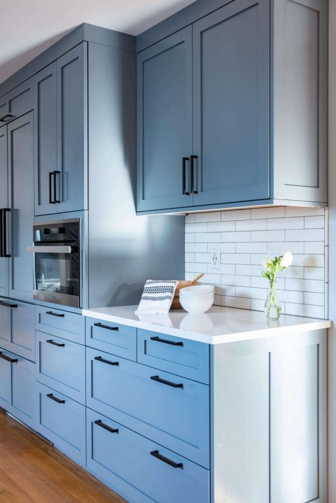 DSC6685-min-scaled Modern kitchen with blue cabinets, black handles, white countertop, and subway tile backsplash—a Serene Family Kitchen Remodel. A bowl with a whisk, wooden spoon, towel, and vase of white flowers grace the counter.