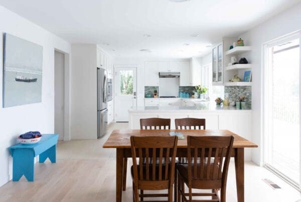 Bright kitchen with white cabinets, stainless steel appliances, a wooden dining table for four, a blue bench, artwork on the wall, and sunlight streaming in—perfect for a West Seattle Artist Retreat.