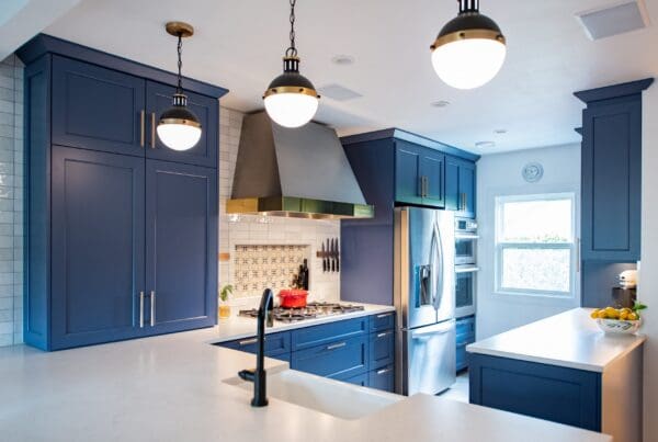 Modern Tudor kitchen remodel with blue cabinets, stainless steel appliances, white countertops, pendant lights, a gas stove with a red pot, and a window letting in natural light. Lemons sit in a bowl on the right counter.