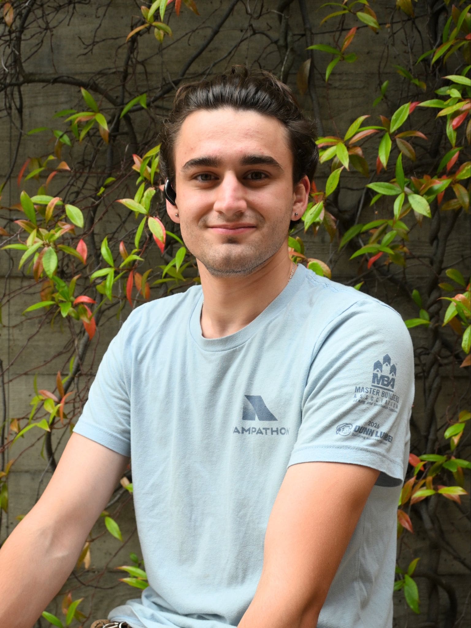 Blake Brannon Blake Brannon headshot. Blake is wearing a light blue T-shirt sits outdoors in front of a vine-covered wall, smiling softly at the camera.