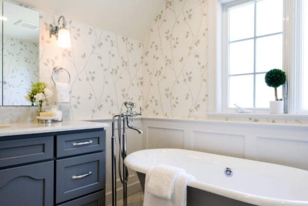 Elegant bathroom with freestanding clawfoot tub, gray vanity, light floral wallpaper, and window. A white towel drapes over the tub—a perfect touch for a Romantic Bathroom Remodel. Potted plant rests on the windowsill.