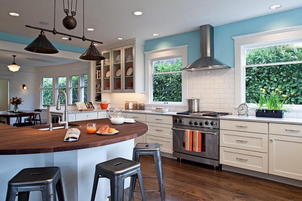 MG_5737-1 Modern kitchen with white cabinets, stainless steel appliances, a round wooden island with stools, bright blue walls, and large windows showing green foliage—a stunning Queen Anne Kitchen Remodel. Fruit and drinks sit on the island.