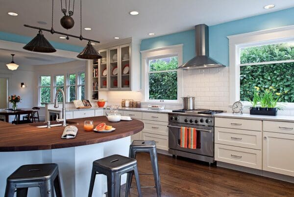 Modern kitchen with white cabinets, stainless steel appliances, a round wooden island with stools, bright blue walls, and large windows showing green foliage—a stunning Queen Anne Kitchen Remodel. Fruit and drinks sit on the island.