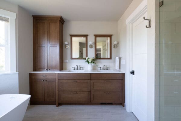 Modern bathroom from a sophisticated Seattle Second Story Remodel with a double vanity featuring wooden cabinets, two mirrors, and white sinks. A vase with flowers sits between the sinks; tub and glass shower are partially visible.